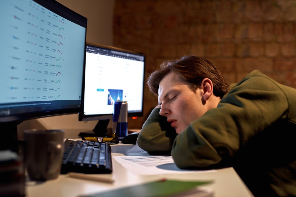 Tired young male trader sleeping at desk with stock market data on computer monitors